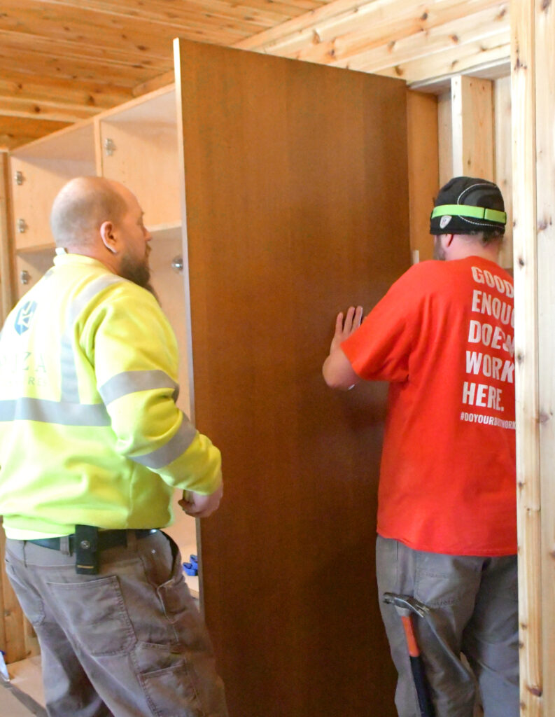 Two men install the side piece of a closet in the entryway of a home.