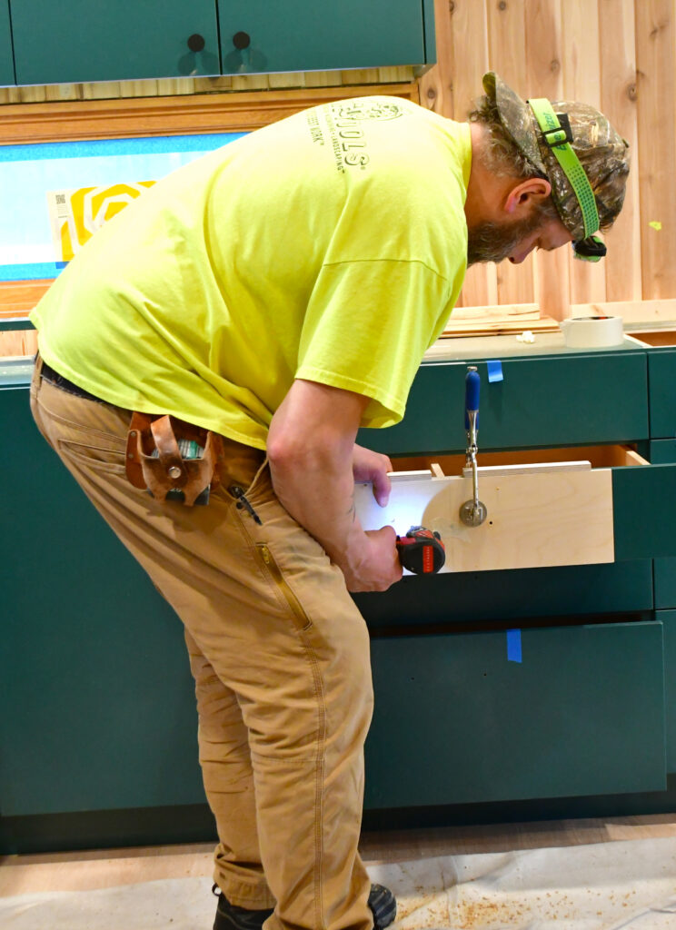 A man uses a wooden jig to pre-drill the holes on a drawer for the drawer pull hardware.