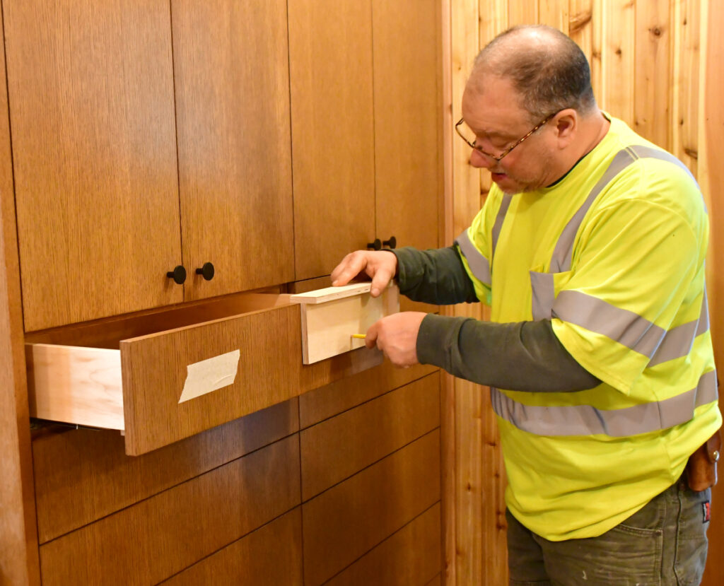 A man uses a wooden jig to find the location to mark where the drawer pull for a cabinet drawer is.